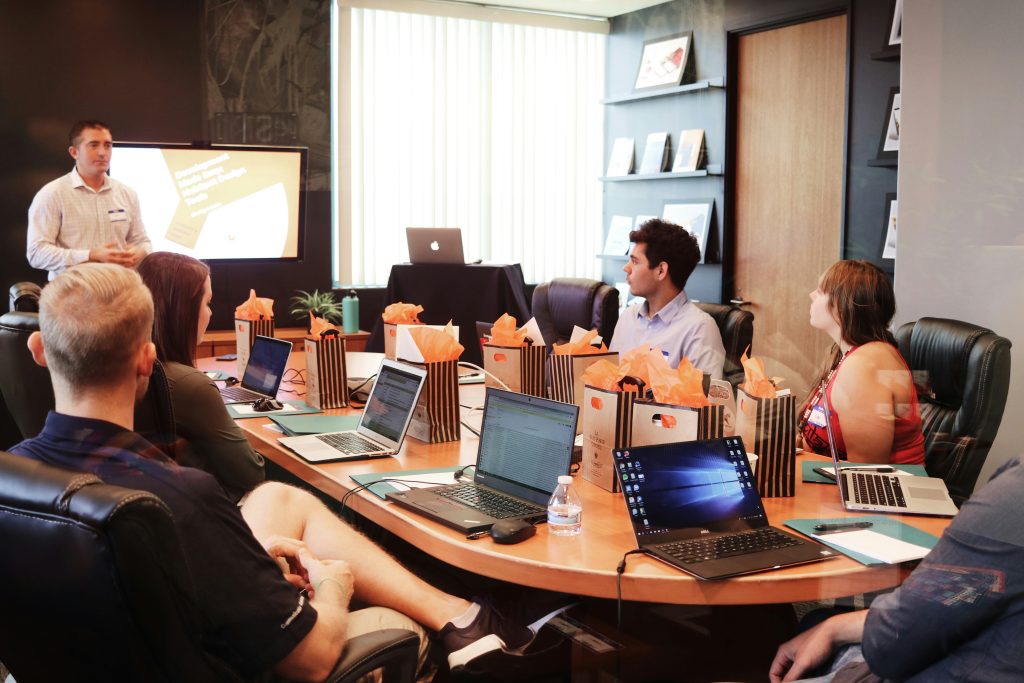 A group of people in a conference room attending a presentation, with laptops open on the table and a presenter standing near a screen displaying slides.