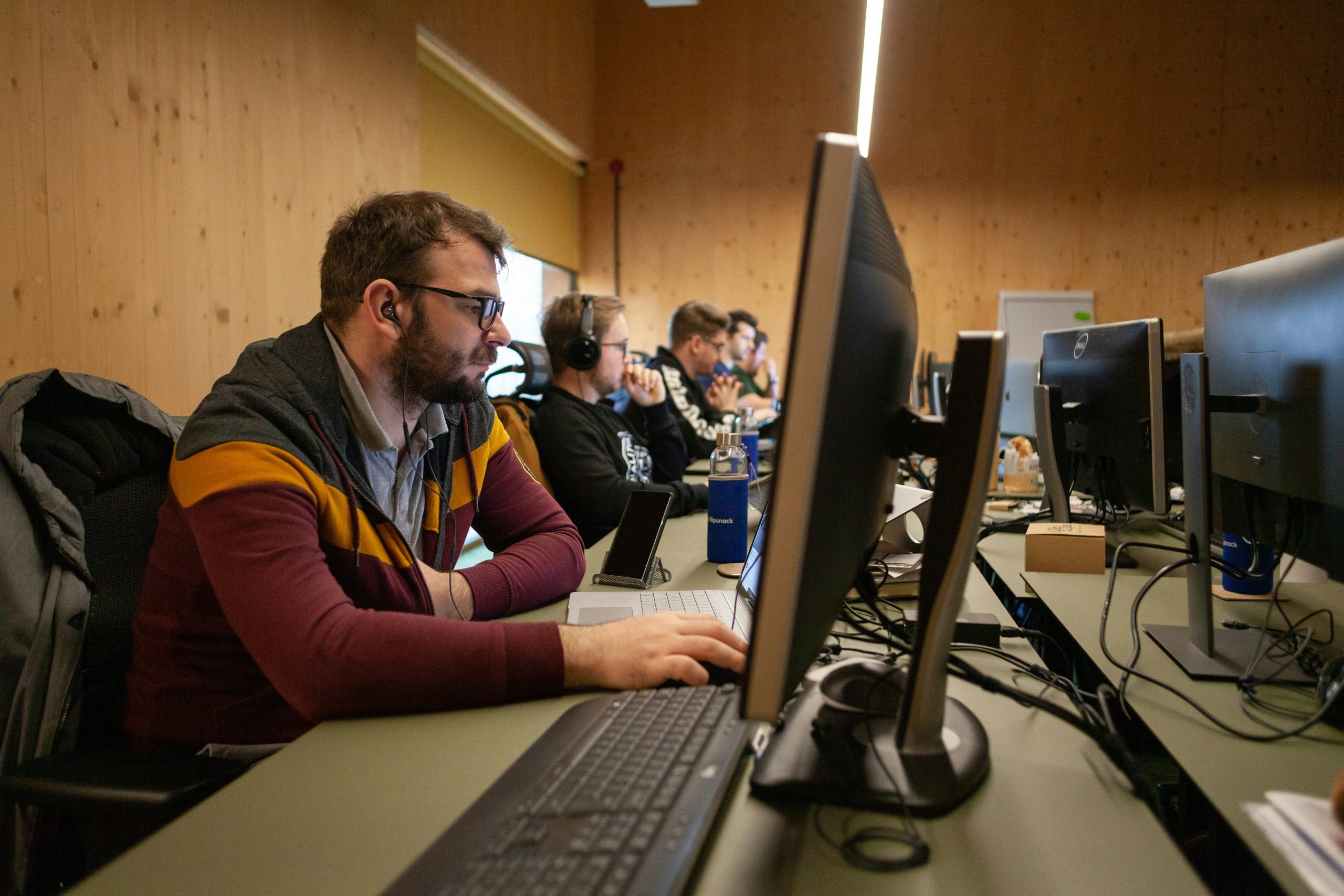 A group of people working at computer desks in an office, each focused on their monitors, with headsets and keyboards in front of them.
