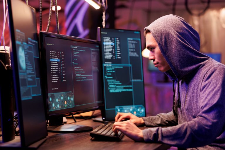 Person in a hoodie working on multiple monitors displaying code and security data.