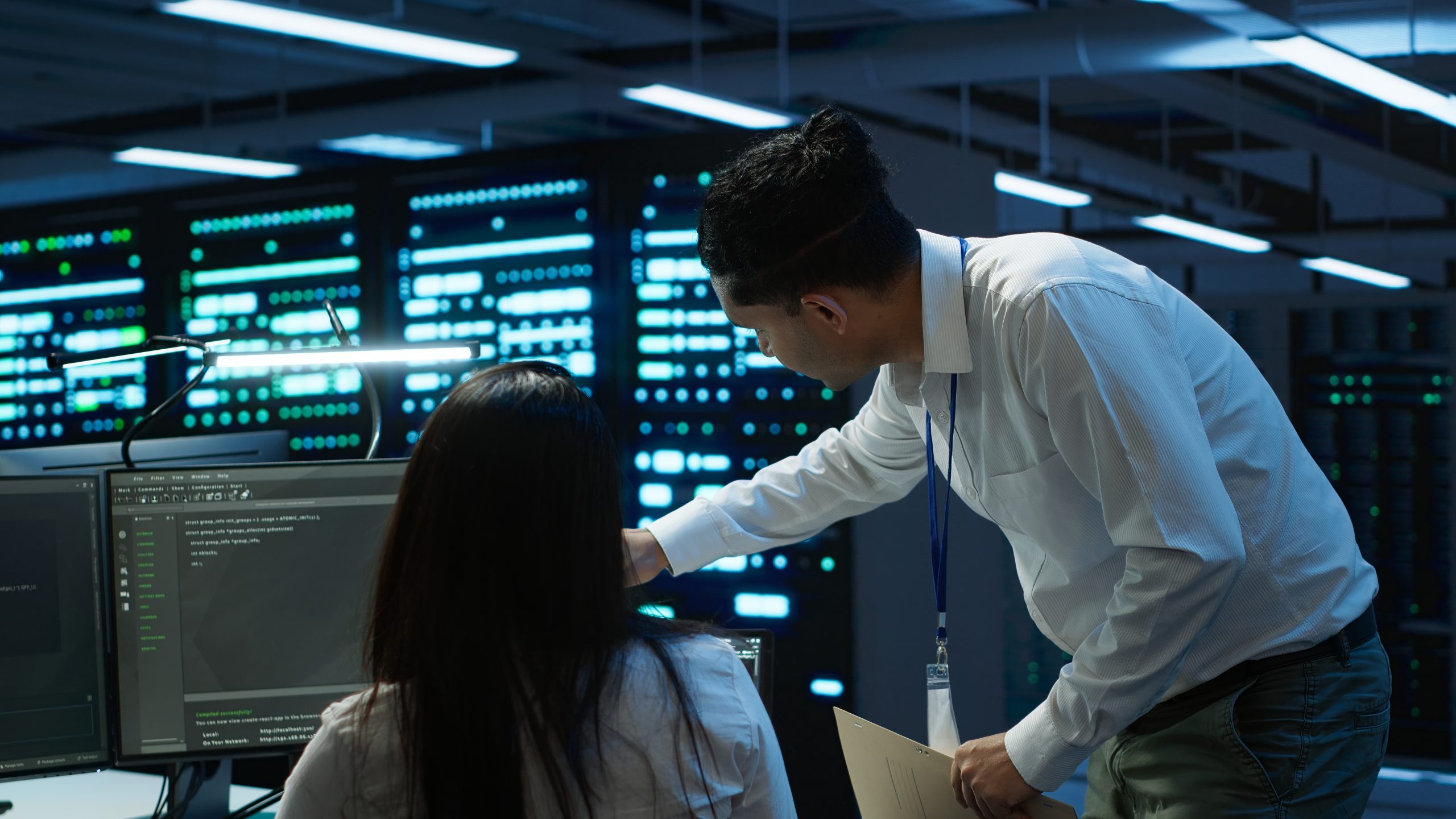 Engineer guiding colleague while reviewing code on monitors.